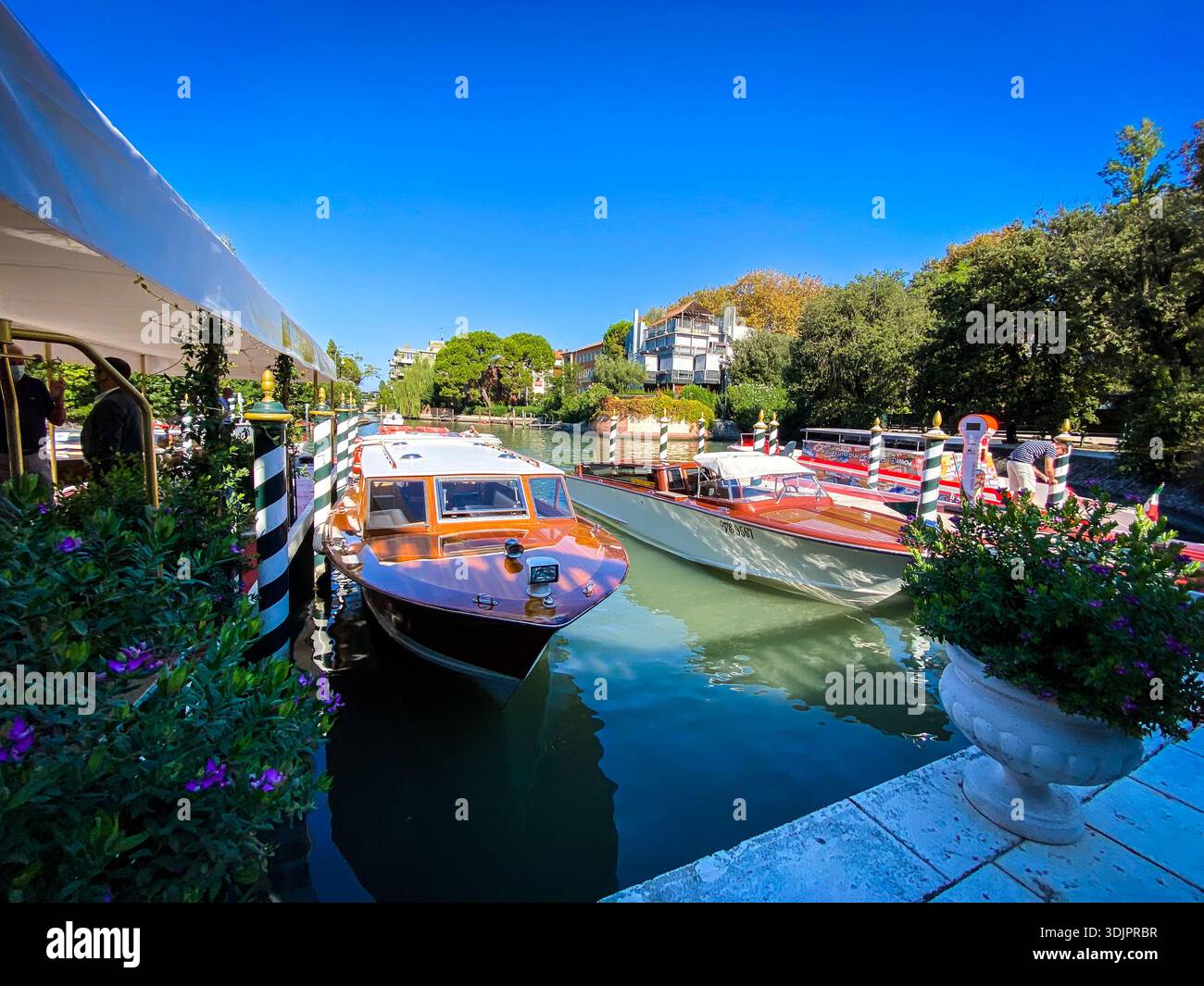 Bateaux à moteur en bois orange classiques amarrés à des poteaux rayés le long d'un canal serein de Venise, avec terrasse au bord de l'eau, grands pots de fleurs et pins sous-jacents - Image de stock capturée avec un smartphone