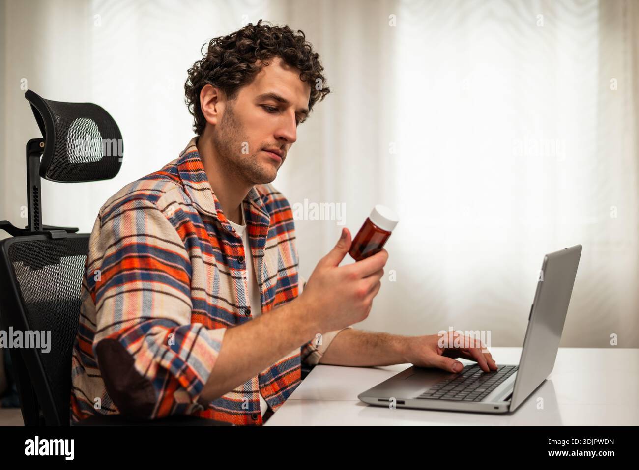 Jeune homme utilisant un ordinateur portable pour rechercher des détails sur ses médicaments prescrits tout en étant assis à sa maison confortable. Banque D'Images