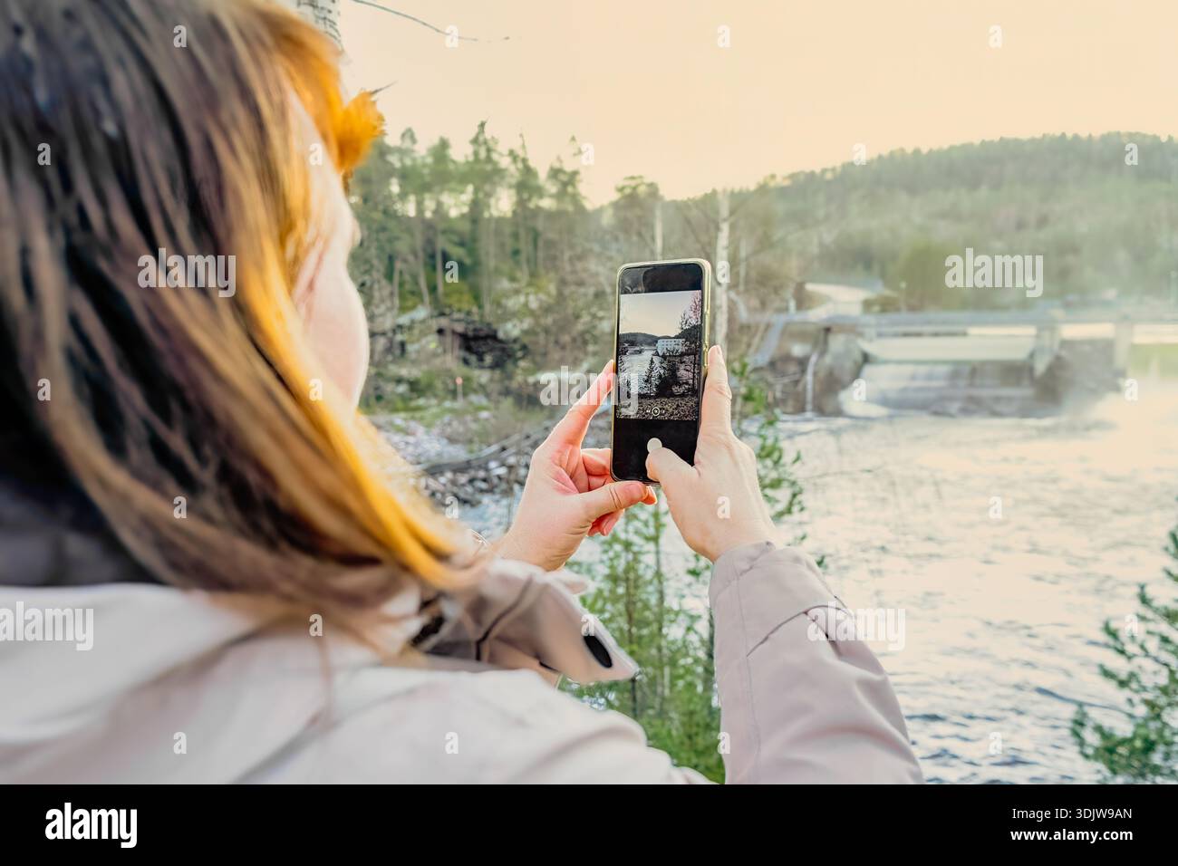 Photographe femme prend des photos de la rivière et du barrage avec smartphone, immergé dans la nature, entouré d'arbres et de paysages vibrants, profitant de la publicité en plein air Banque D'Images