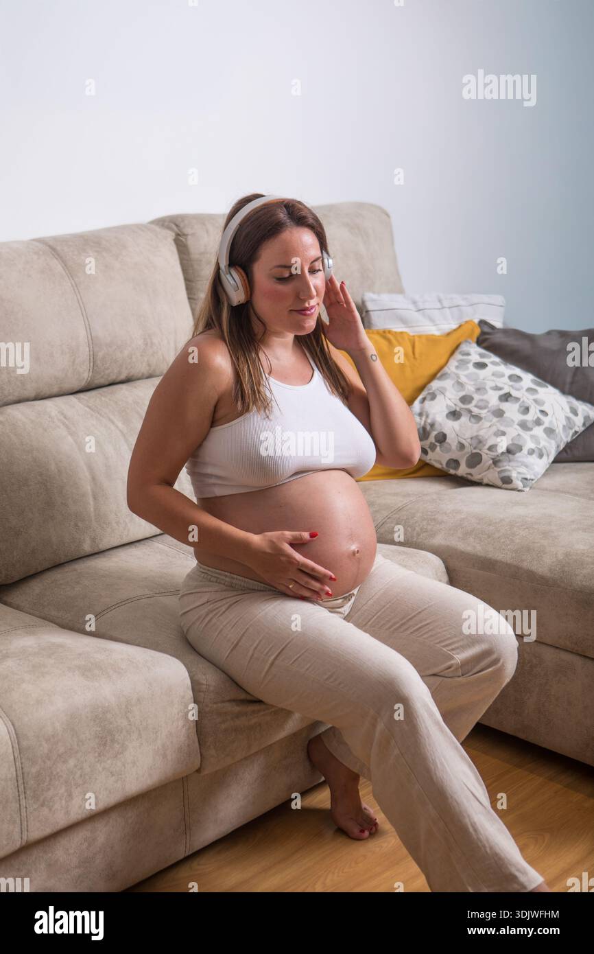 Une femme enceinte s'assoit confortablement sur son canapé, écoutant de la musique douce à travers des écouteurs. Elle berce doucement sa bosse de bébé tout en profitant du pois Banque D'Images