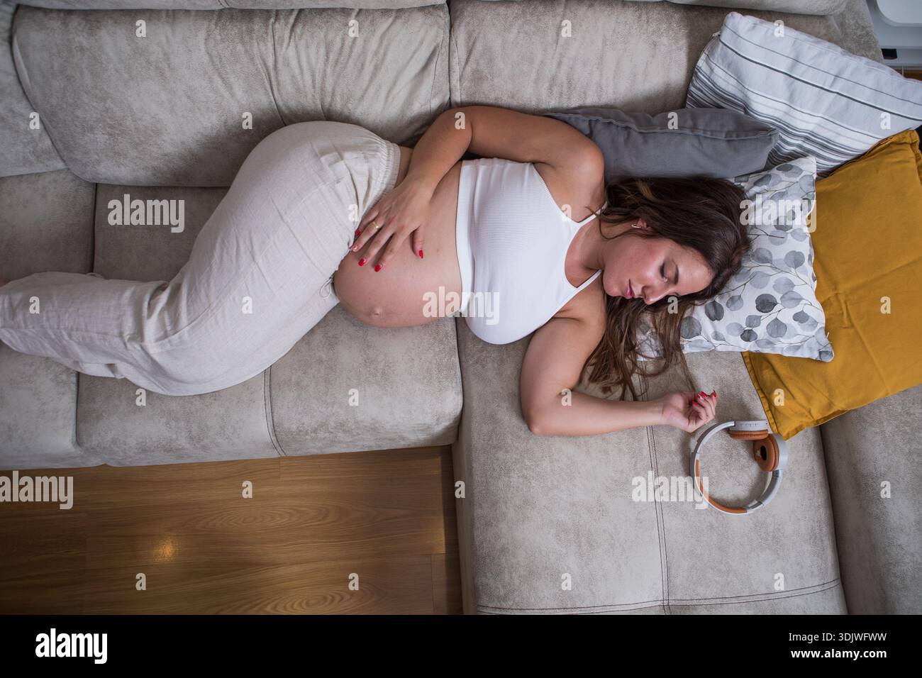 Une femme enceinte se détend sur un canapé élégant, berçant son ventre doucement. Des oreillers moelleux l'entourent comme elle profite d'un moment de calme dans un espace de vie confortable Banque D'Images