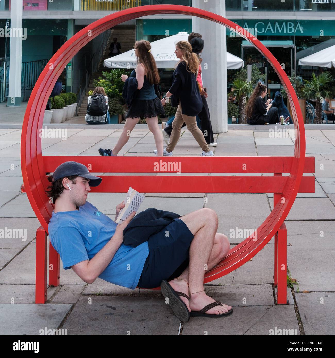 Un jeune homme se détend en lisant sur un banc rouge circulaire à la place publique urbaine de London South Bank tandis que les piétons passent devant. Banque D'Images