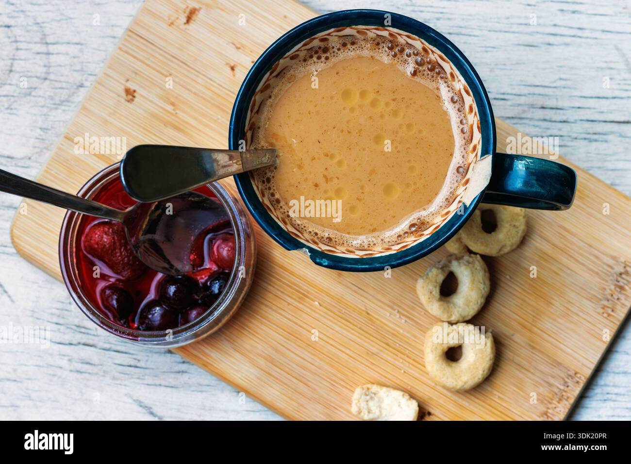 Vue de dessus d'une tasse à café sur une planche de bois avec des biscuits, du beurre, et un pot ouvert de confiture de fruits sauvages avec une cuillère, sur une table légère avec espace de copie. Banque D'Images
