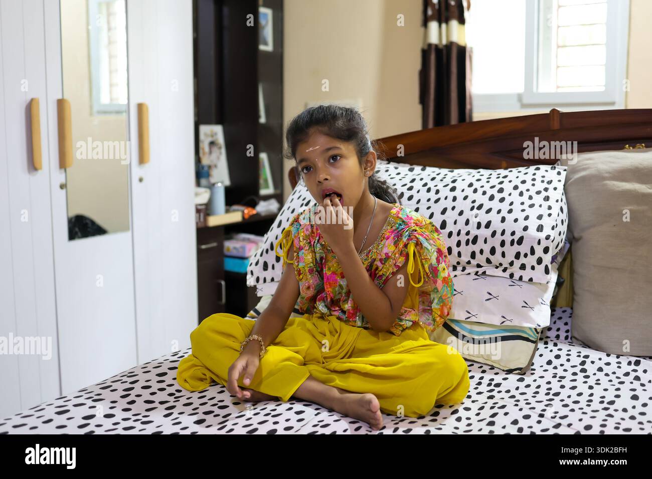 Un enfant est assis sur un lit et mange une collation tout en regardant autour de l'intérieur dans un cadre familial. Banque D'Images