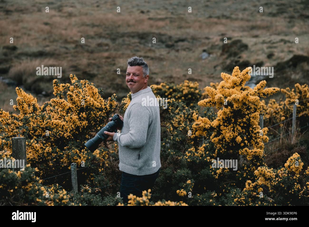 Photographe debout parmi les arbustes à fleurs jaunes dans un paysage naturel, tenant un téléobjectif Banque D'Images