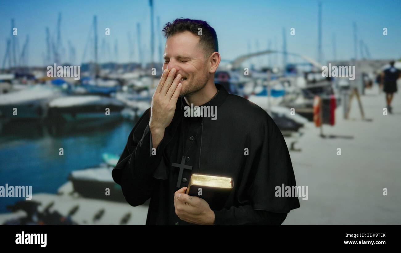 Homme souriant en tenue de prêtre tenant la Bible dans une marina avec des bateaux en arrière-plan, véhiculant un sentiment de contemplation et de joie au bord de la mer. Banque D'Images