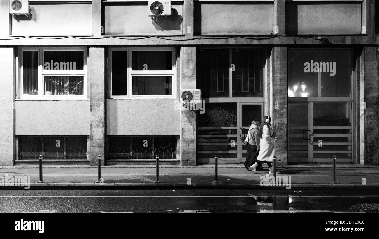 Deux piétons marchant ensemble le long d'un trottoir urbain devant un bâtiment moderne en béton, créant un paysage de photographie de rue calme et intemporel Banque D'Images