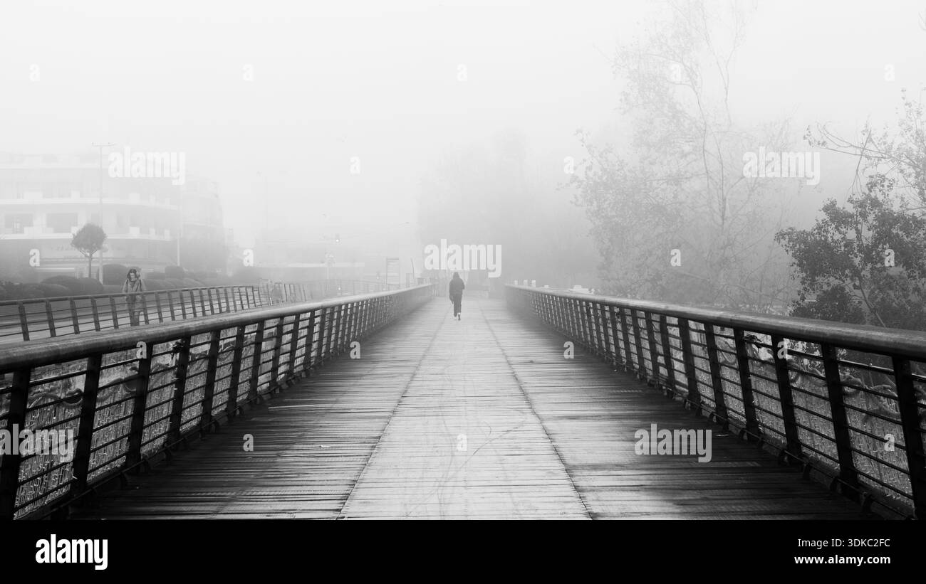 Personne solitaire traversant un pont piétonnier dans un brouillard dense. Scène urbaine en noir et blanc avec une forte symétrie et profondeur. Banque D'Images