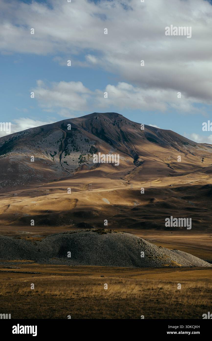 Montagne ensoleillée s'élevant au-dessus d'un plateau doré sec, avec des nuages spectaculaires et des ombres douces formant un vaste paysage intact. Banque D'Images