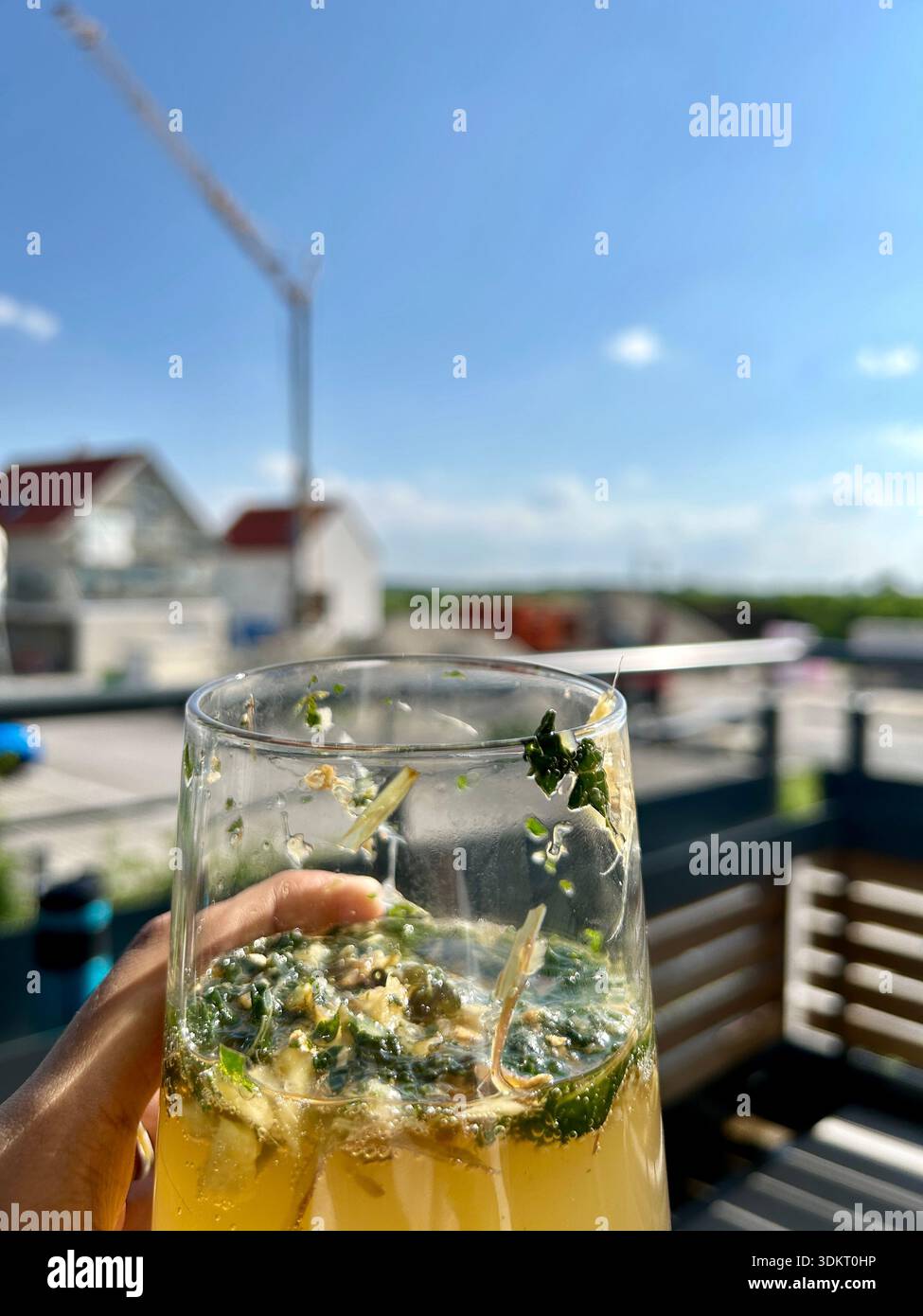 Gros plan d'une main tenant un verre de cocktail aux herbes fraîches (Hugo Spritz ou Mojito) sur un balcon ensoleillé. Fond urbain avec ciel bleu Banque D'Images