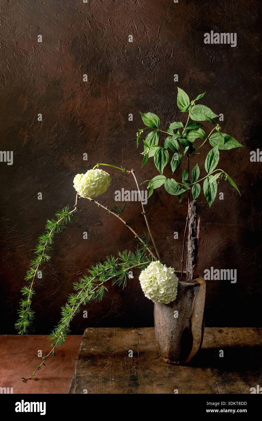 Nature morte artistique avec ikebana fleurs d'hortensia blanc feuillage vert et branches de pin dans un vase en bois vintage sur fond texturé foncé. Eleg Banque D'Images
