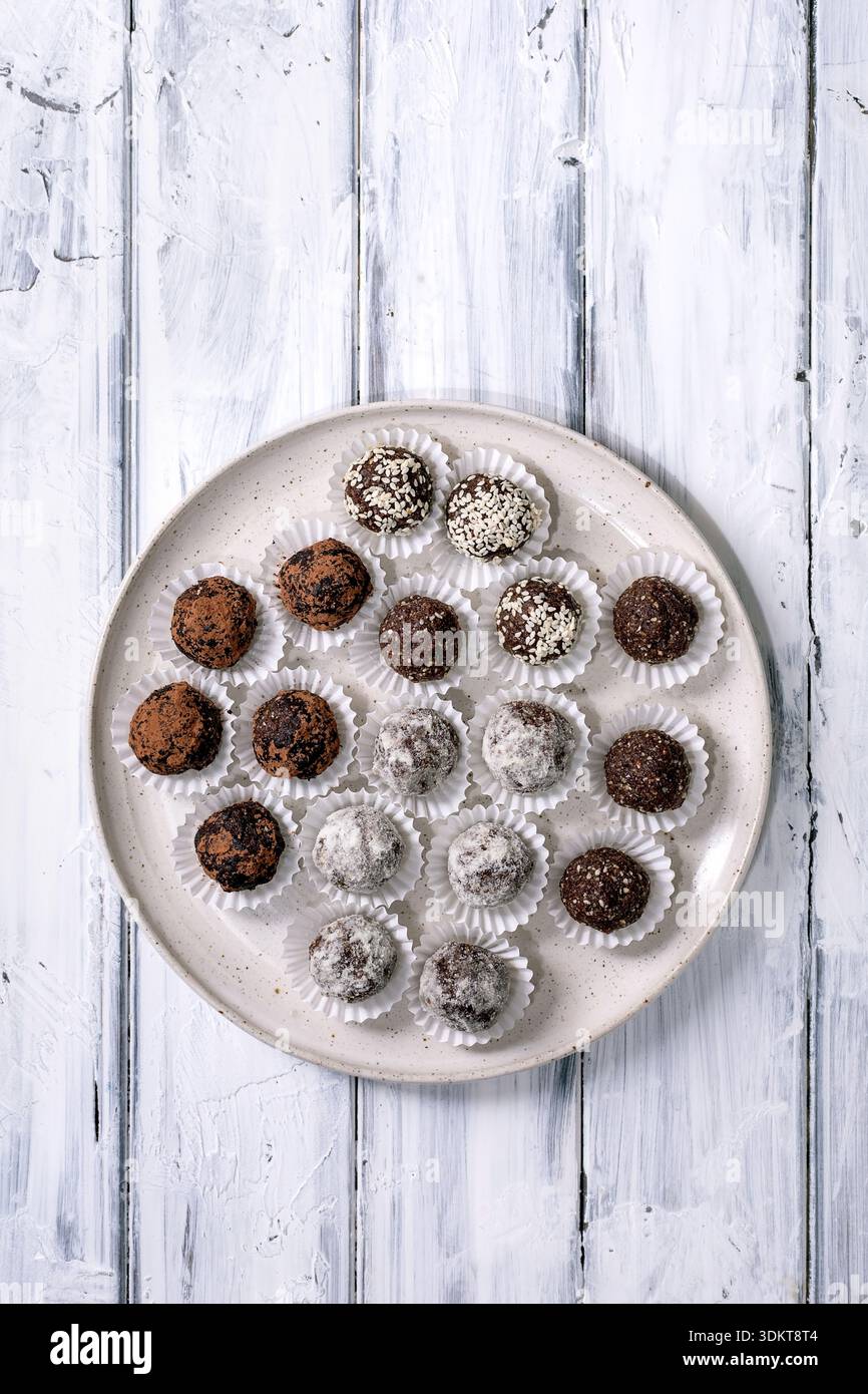 Assortiment de boules végétaliennes crues truffes au chocolat maison avec différentes garnitures sucre en poudre cacao en poudre graines de sésame sur plaque en céramique. Blanc en bois b Banque D'Images