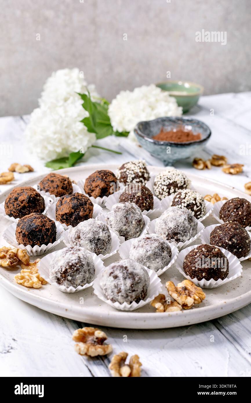 Assortiment boules végétaliennes crues au chocolat maison truffes avec différentes garnitures sur assiette blanche. Noix de cacao en poudre dispersées autour. Petits bols avec Banque D'Images