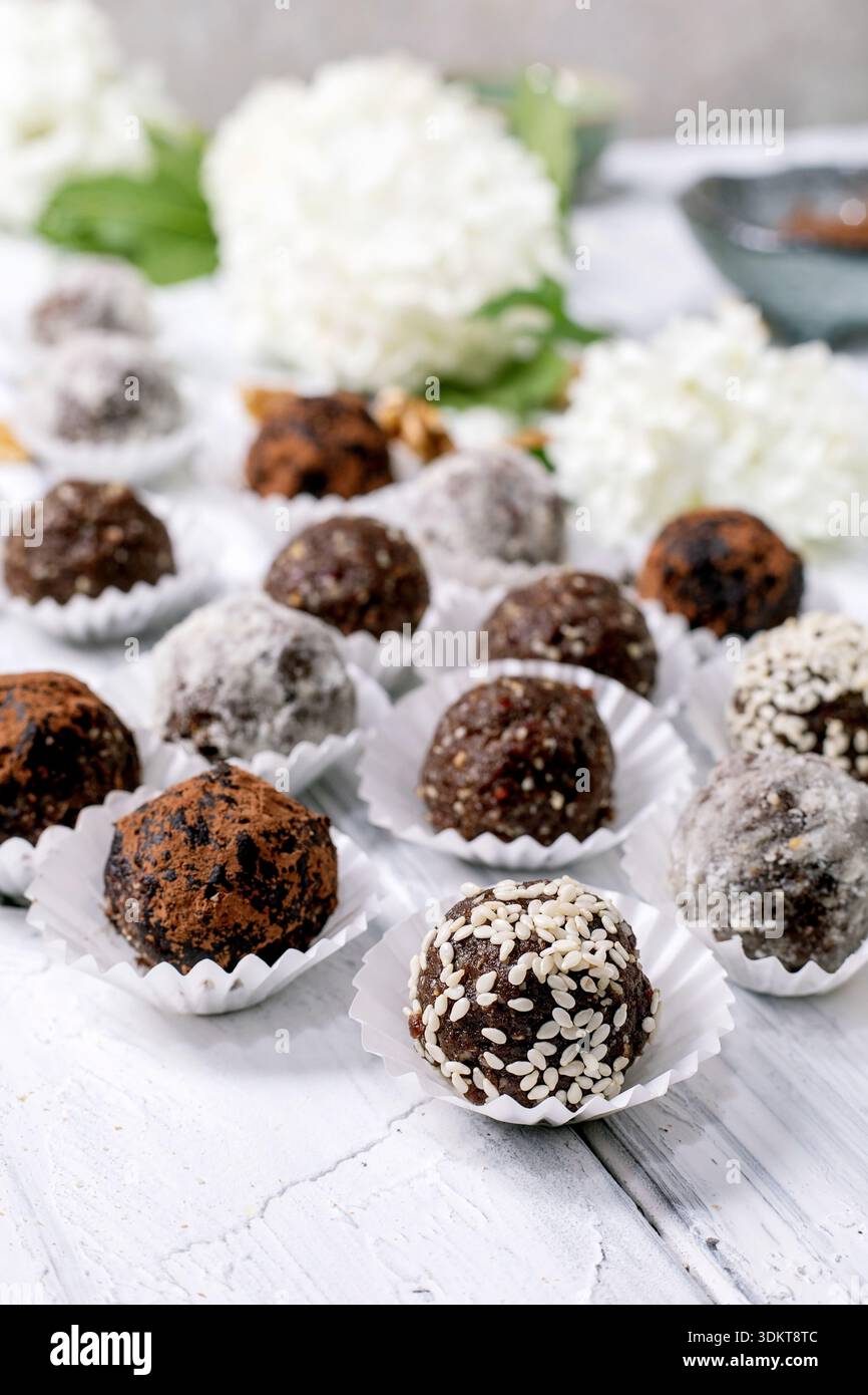 Divers boules végétaliennes crues au chocolat truffes, enrobées de poudre de cacao graines de sésame sucre en poudre placé dans des gobelets en papier sur une table en bois blanc avec hydra Banque D'Images