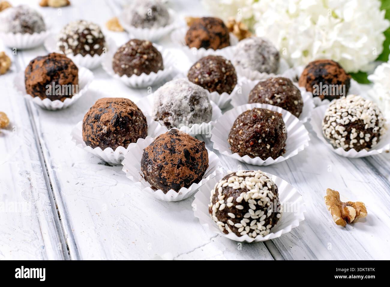 Assortiment de boules végétaliennes crues au chocolat maison truffes avec différentes garnitures dans des gobelets en papier sur fond de bois blanc décoré de fleurs blanches Banque D'Images