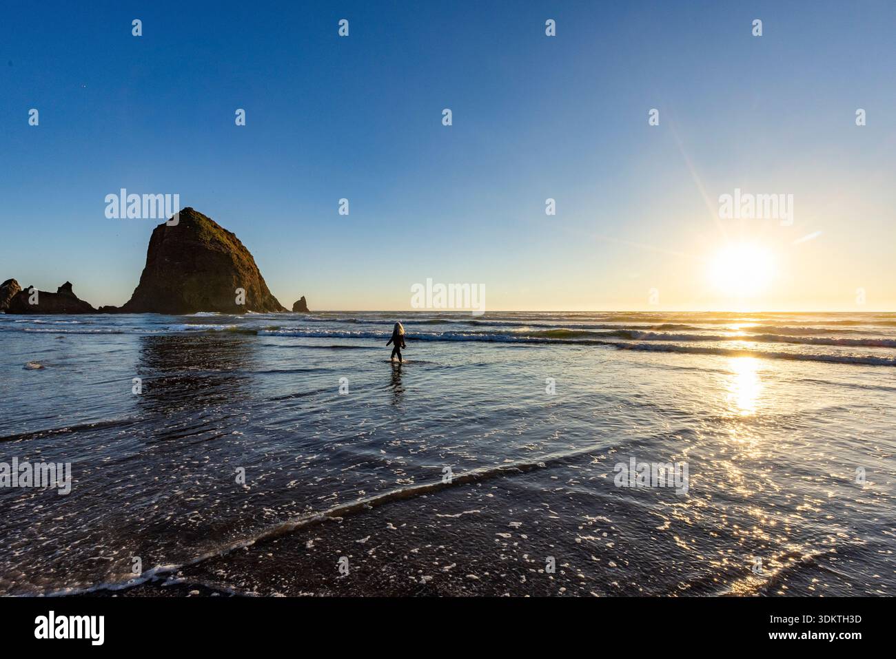 USA, Oregon, Cannon Beach, femme pataugant dans le surf près du rocher de meule de foin au coucher du soleil Banque D'Images