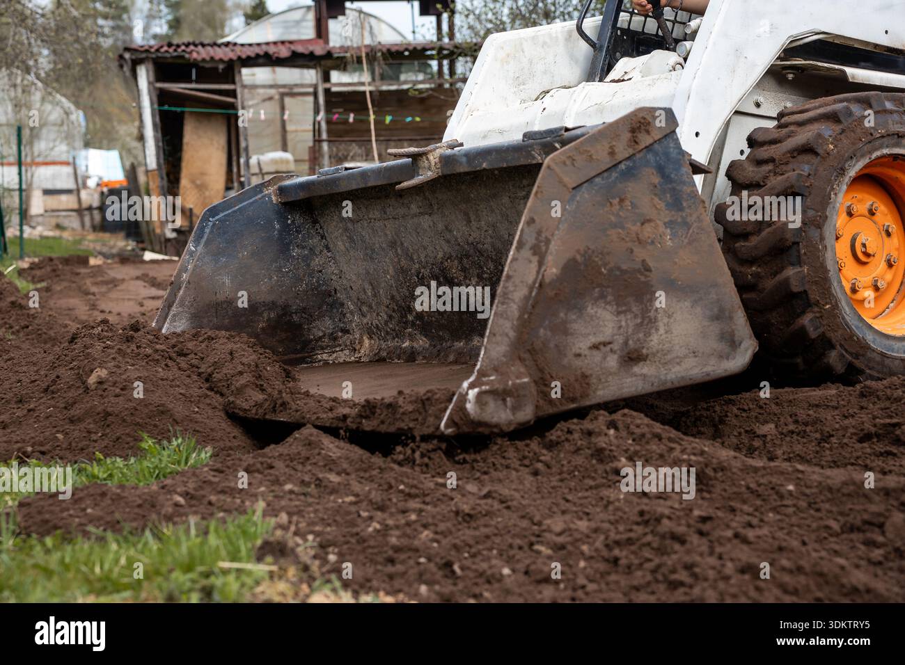 Un petit véhicule de chantier avec une roue orange et un chargeur frontal déplace le sol près d'une structure rustique avec un toit en métal ondulé et des panneaux en bois. Banque D'Images