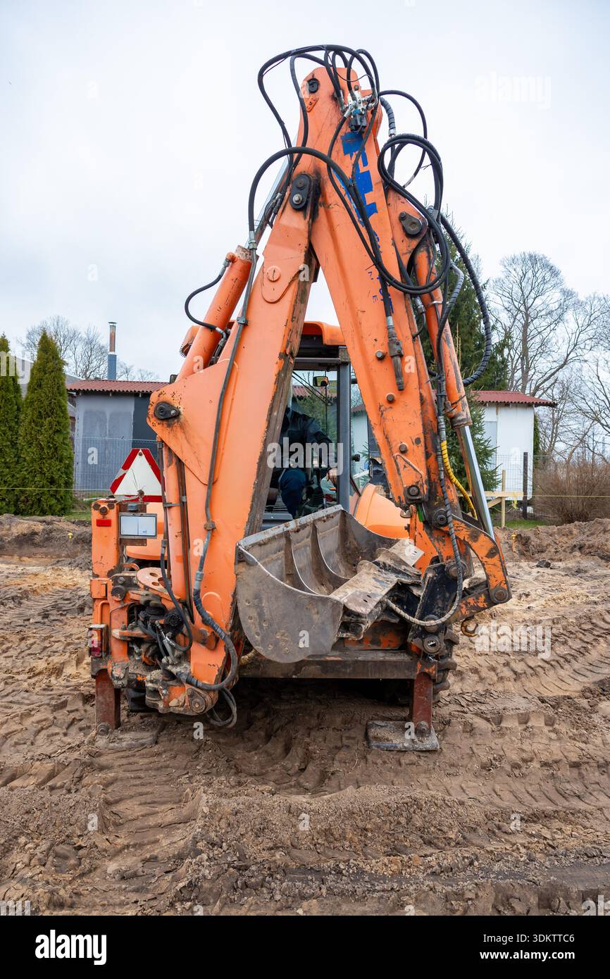 Une pelle orange avec flexibles hydrauliques visibles est installée sur un chantier de construction. Un bâtiment blanc avec un toit rouge est entouré d'arbres et d'une clôture. Banque D'Images