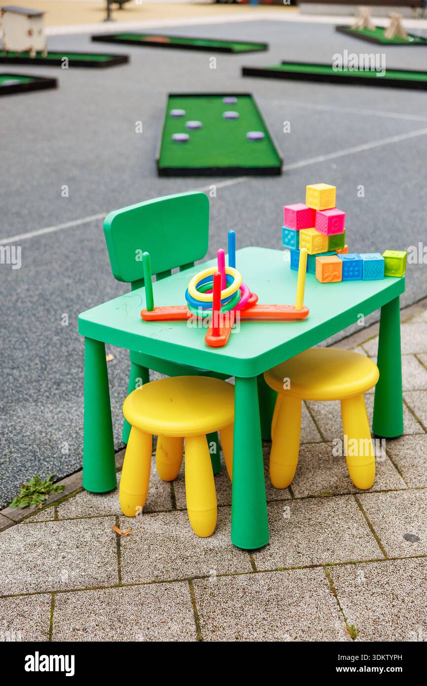 Une petite table verte, une chaise et deux tabourets jaunes tiennent des anneaux et des blocs souples à côté de chemins pavés près de verts de putting compacts pendant la journée, avec cri Banque D'Images
