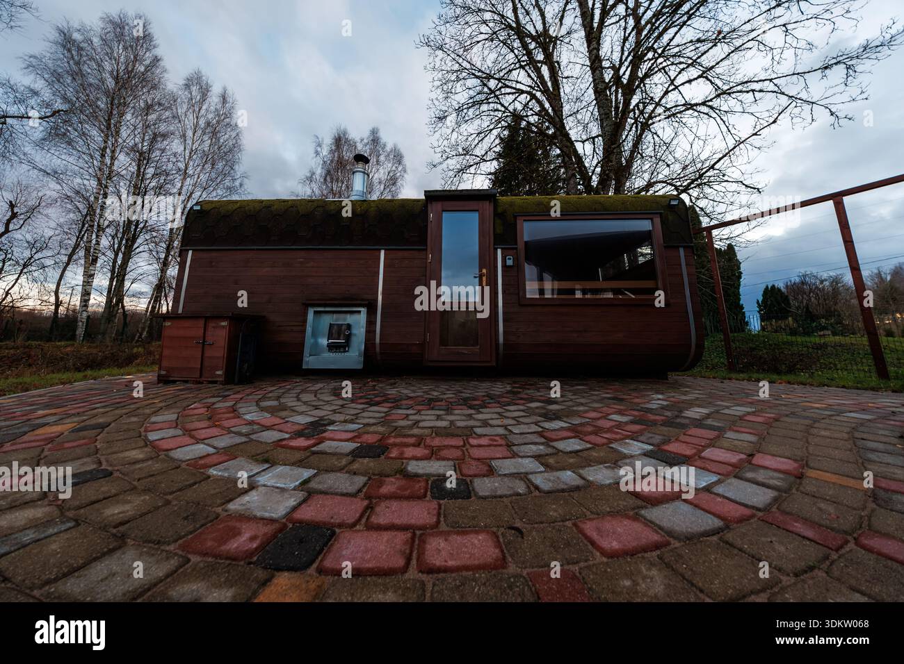 Une cabine en bois en forme de tonneau avec des bardeaux moussseux se trouve sur des pavés en spirale multicolores, la fenêtre et la porte en verre réfléchissent les branches de bouleau, la lumière diffuse fraîche s. Banque D'Images