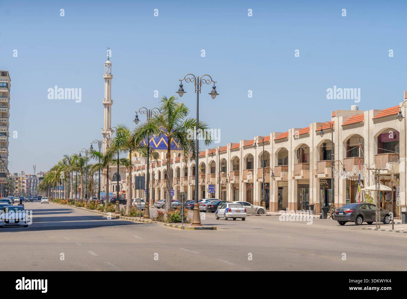 Port Said boulevard avec mosquée, minaret, palmiers, lampes ornées et bâtiments beiges voûtés le long de l'avenue de la circulation. Banque D'Images