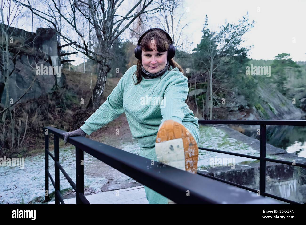 Athlète féminine effectue l'étirement des jambes sur la balustrade dans un endroit extérieur tranquille, habillé dans une tenue verte chaude, mettant l'accent sur la forme physique et le dévouement à la guérison Banque D'Images