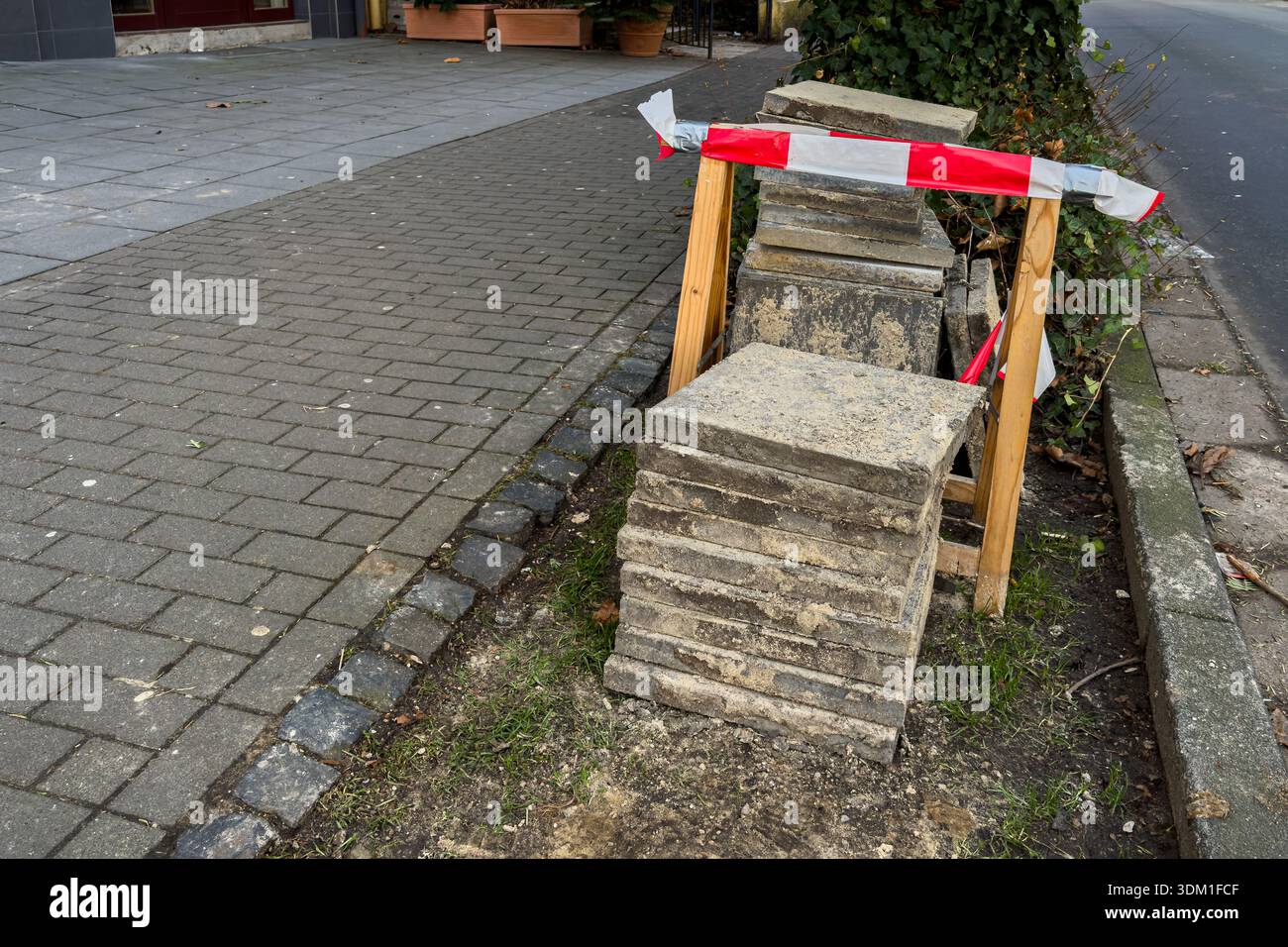 Les travailleurs font des travaux de construction sur le trottoir où des blocs sont empilés près d'un bâtiment. Du ruban rouge et blanc est placé autour de la zone de travail pour avertir Banque D'Images