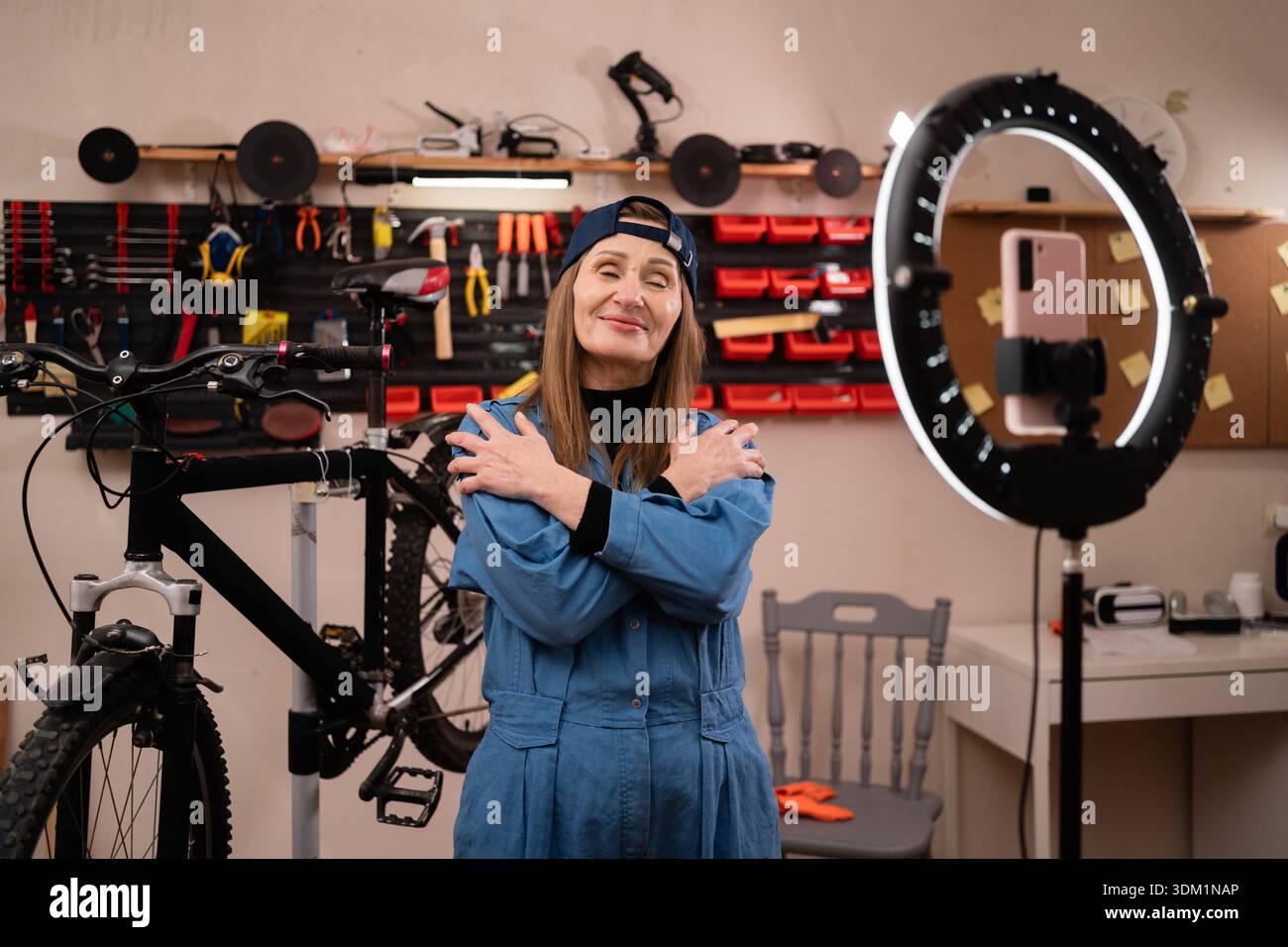 Souriant femme heureuse blogueuse influencer streaming en direct travaillant dans le garage, vlogger s'embrassant les yeux fermés sur la caméra pour examiner. mécanicien de vélo en s. Banque D'Images