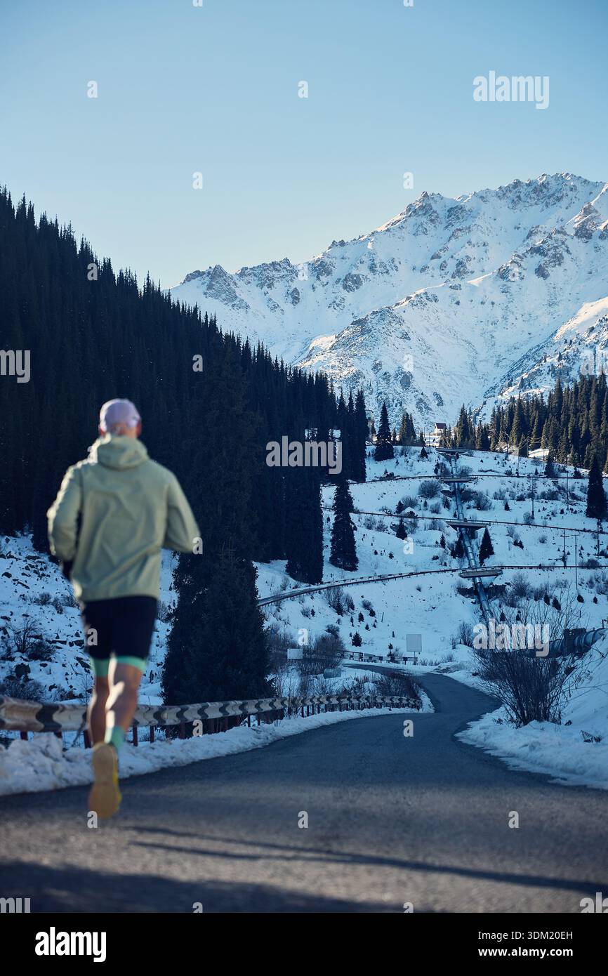 Homme athlète courir sur la route dans la vallée de la montagne de neige à la journée ensoleillée d'hiver, hors de portée. Jogging en vêtements de sport à la nature. Banque D'Images