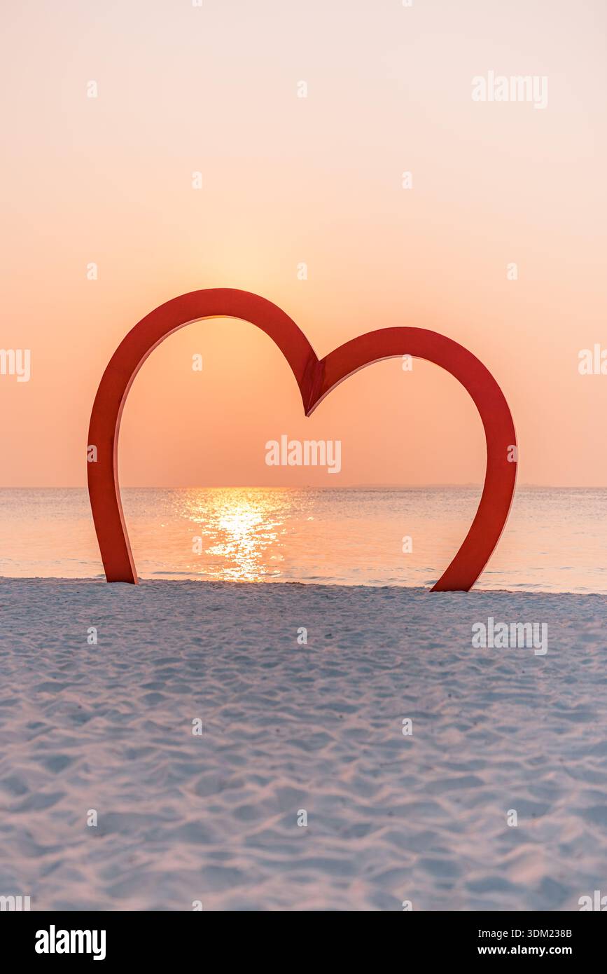 Symbole d'amour en forme de cœur rouge debout sur le sable d'une plage tropicale à un coucher de soleil doré au bord de la mer. Lueur romantique du ciel rose. Voyage de noces anniversaire de couple vacances Banque D'Images
