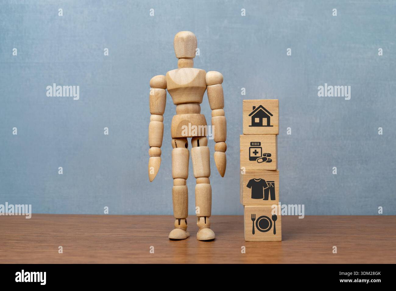 Une poupée en bois se tient à côté de blocs empilés avec des icônes pour le logement, les soins de santé, les vêtements et la nourriture. Besoins humains fondamentaux, coût de la vie Banque D'Images