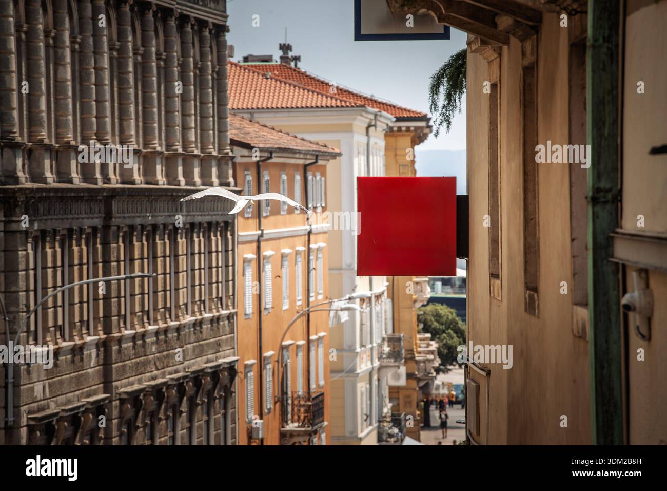 Panneau carré rouge blanc suspendu au-dessus d'une rue étroite dans une ville de style méditerranéen, offrant un espace de copie pour l'image de marque ou la publicité, avec un bâtiment ancien Banque D'Images