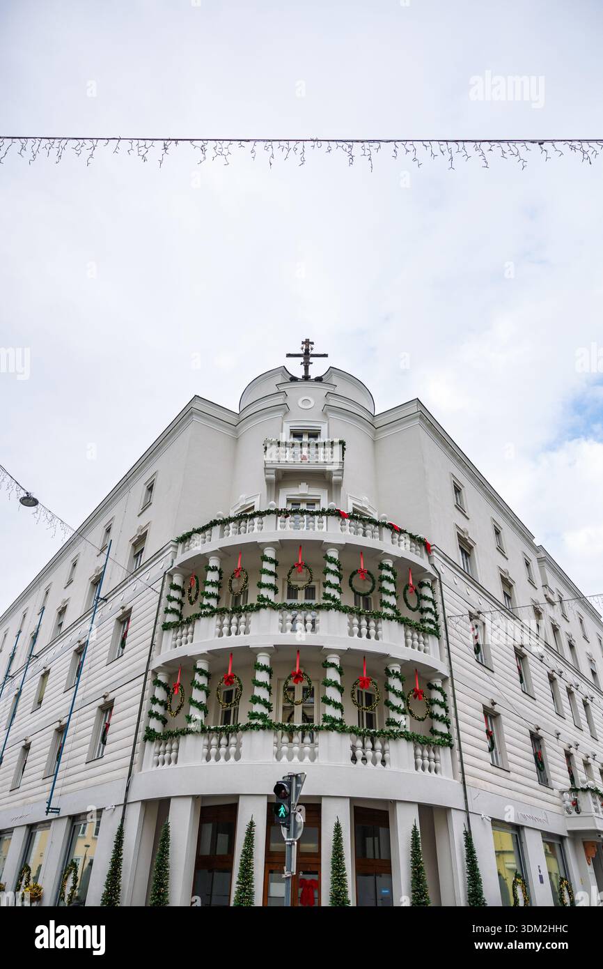 Un charmant bâtiment d'angle blanc drapé de guirlandes vertes et de rubans rouges, avec balcons et couronnes. Devantures de magasins au niveau de la rue et pavés enneigés Banque D'Images