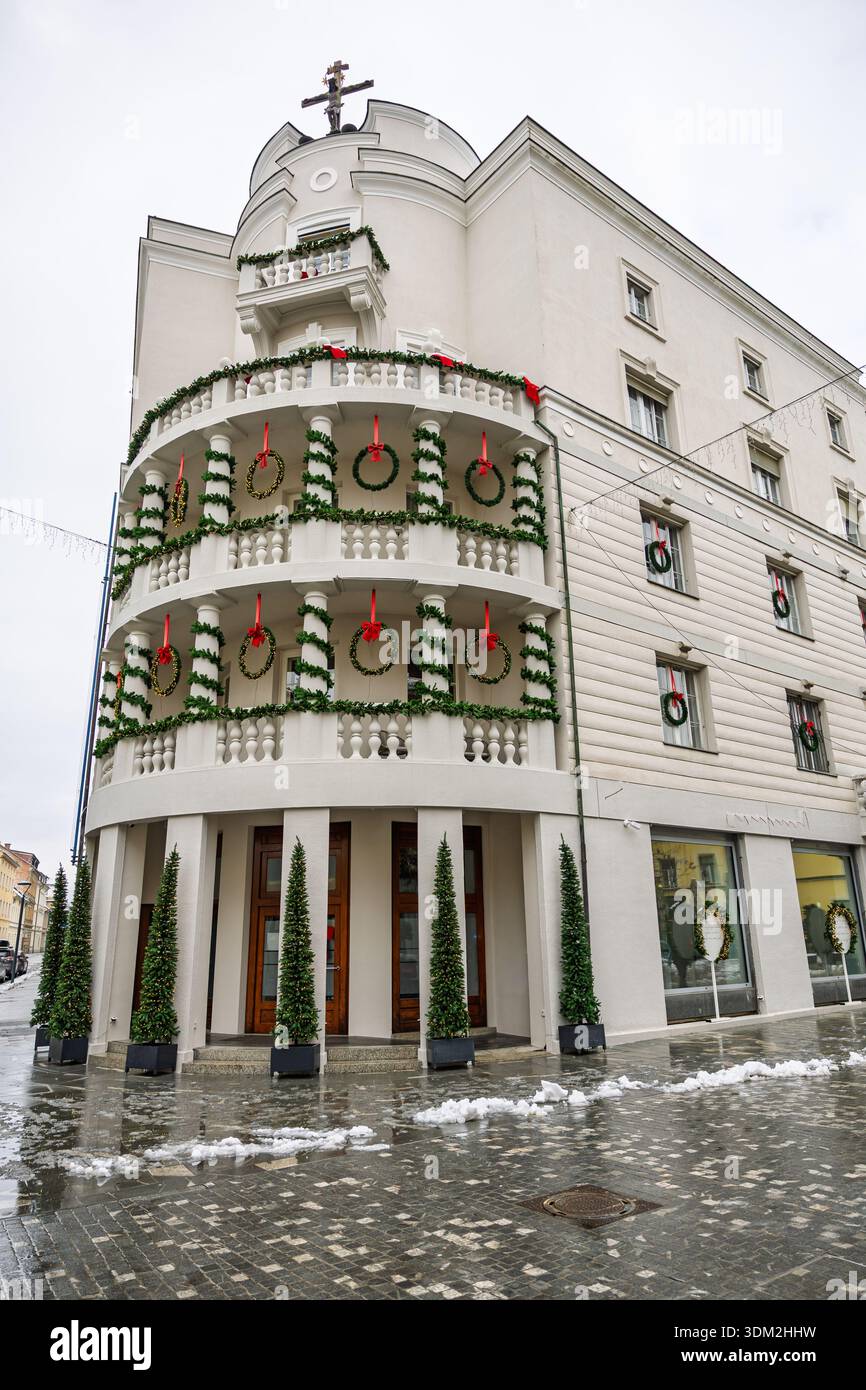 Un charmant bâtiment d'angle blanc drapé de guirlandes vertes et de rubans rouges, avec balcons et couronnes. Devantures de magasins au niveau de la rue et pavés enneigés Banque D'Images