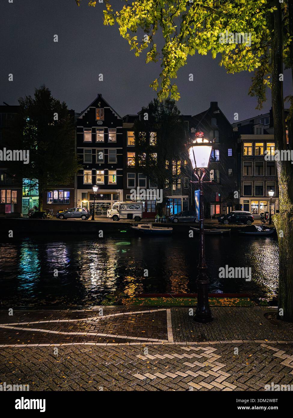 Vue nocturne sur le canal d'Amsterdam bordé de maisons hollandaises à pignons illuminées, lampadaire classique qui brille sous les feuilles jaunes d'automne, reflets en c. - Image de stock capturée avec un smartphone