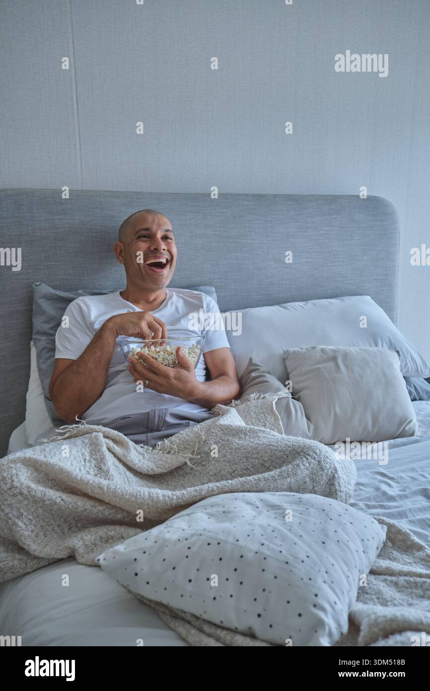 Homme riant tenant un bol de pop-corn appréciant le divertissement dans le lit la nuit de cinéma Banque D'Images