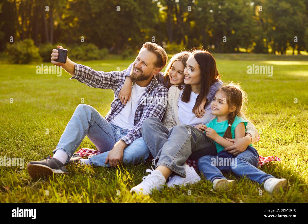 Heureux parents avec deux enfants assis sur l'herbe dans un parc verdoyant ensoleillé et prenant selfie ensemble. Banque D'Images