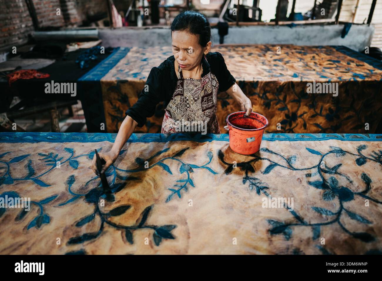 Femme peint des motifs floraux sur tissu dans un atelier de batik, en utilisant la teinture d'un seau pour créer des textiles traditionnels Banque D'Images
