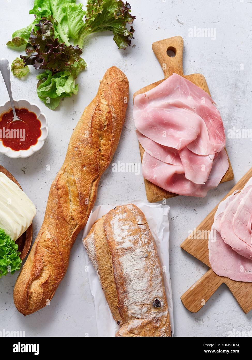 Les ingrédients frais sont disposés sur une table. Une baguette, des tranches de jambon, du fromage mozzarella, de la laitue verte et de la sauce tomate sont prêts. La scène montre un Banque D'Images