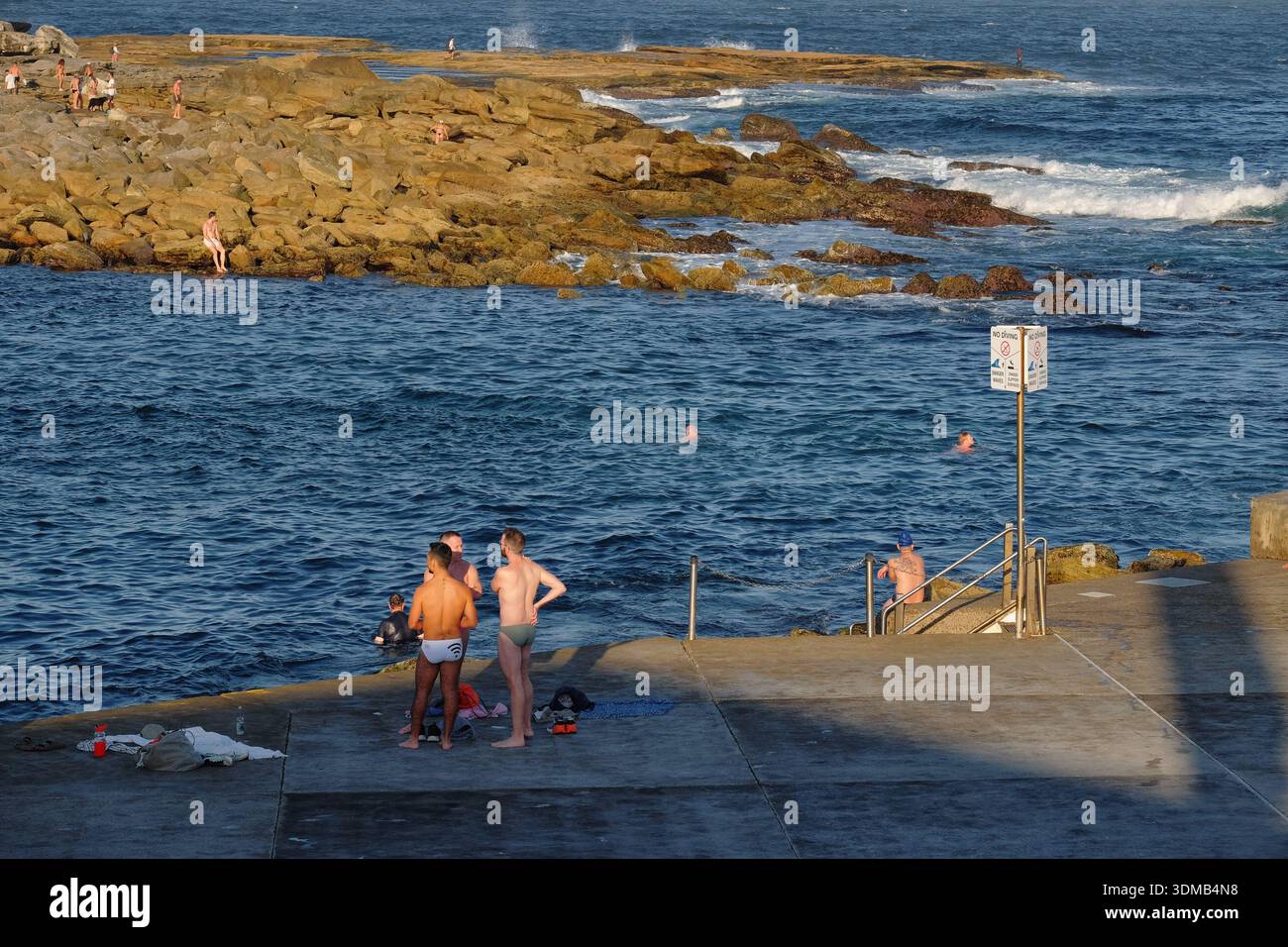 Trois hommes en nageurs debout au soleil sur le bord de la plate-forme avec de l'eau bleu vif de l'océan à Clovelly roche piscine, Sydney, Australie Banque D'Images