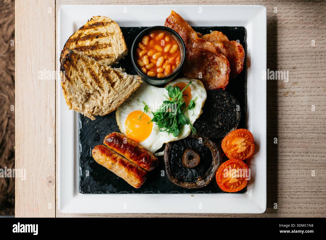 Petit-déjeuner anglais complet traditionnel avec œufs frits saucisses bacon haricots pain grillé et légumes grillés, présentés sur une assiette en ardoise pour les menus du café, Banque D'Images
