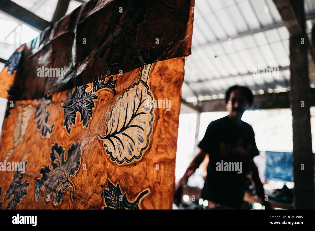 Le tissu batik indonésien avec des motifs floraux audacieux est bien en vue, tandis qu'un artisan flou travaille en arrière-plan d'un atelier en bois rustique Banque D'Images