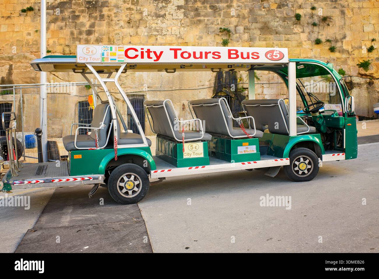 Valletta, Malte - 29 novembre 2025 : vue latérale d'un petit bus vert vide à ciel ouvert électrique de la ville garé dans la rue à côté d'un ancien mur. Banque D'Images