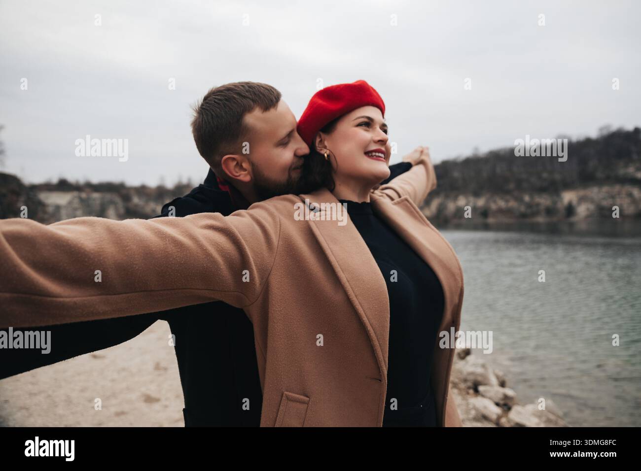 Un homme embrasse une femme avec les bras écartés tout en se tenant au bord de l'eau un jour nuageux. Ils célèbrent leur amour pour la Saint-Valentin. Banque D'Images