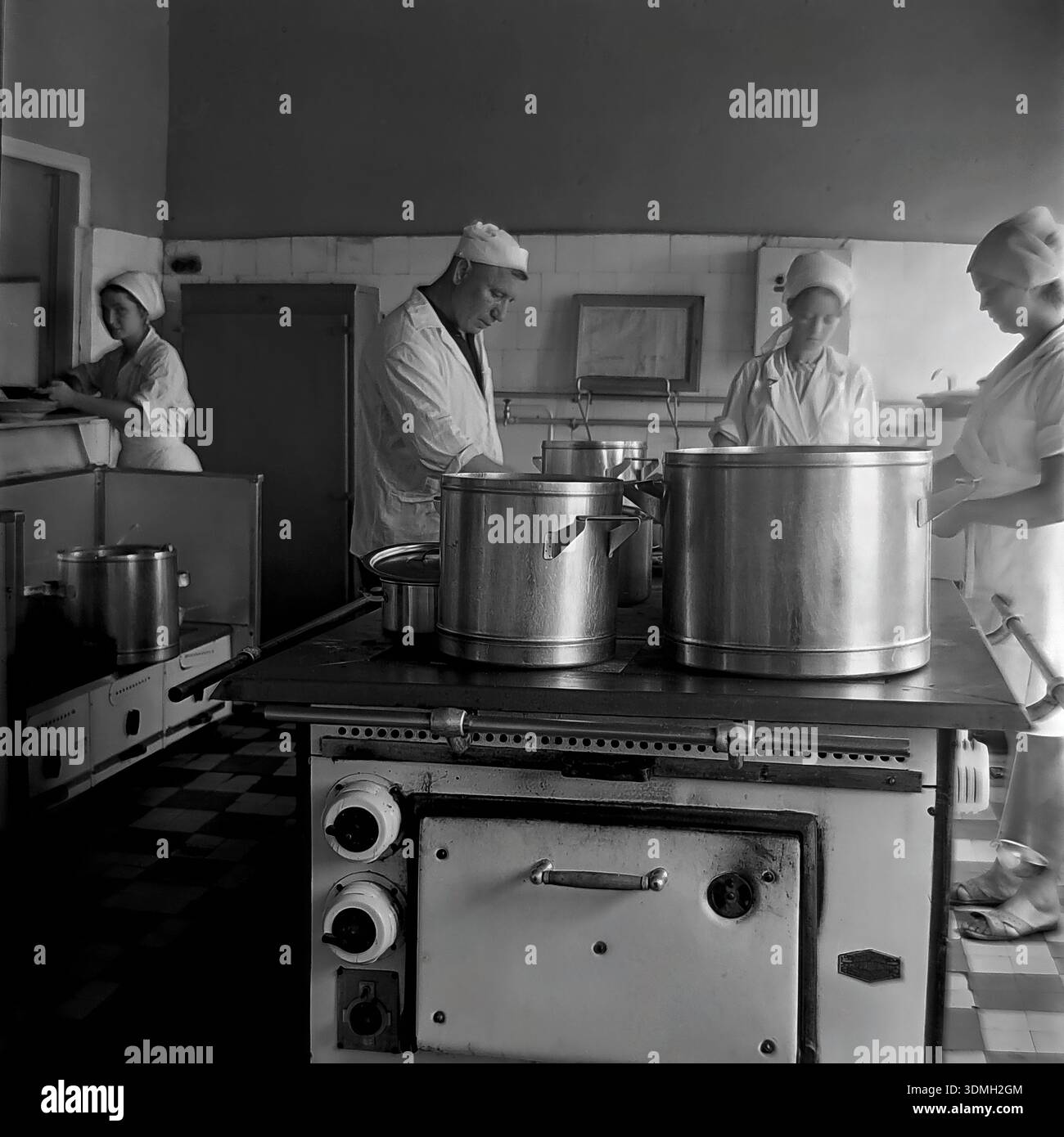 Région de Donetsk, RSS d'Ukraine, Union soviétique, années 1960 Une photo d'archives en noir et blanc capture la cuisine animée d'une cantine de ferme collective locale. Un chef cuisinier masculin et trois assistantes en uniformes sanitaires blancs travaillent dur pour préparer un déjeuner chaud pour les travailleurs de terrain. De grands pots en aluminium mijotent sur un poêle industriel massif dans une pièce carrelée. Cette image vintage documente le système de restauration publique, le travail d'équipe et la vie quotidienne de la paisible région du Donbass pendant l'ère soviétique bien avant la guerre. Banque D'Images