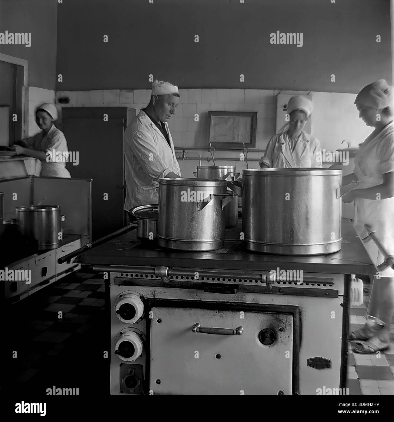 Région de Donetsk, RSS d'Ukraine, Union soviétique, années 1960 Une photo d'archives en noir et blanc capture la cuisine animée d'une cantine de ferme collective locale. Un chef cuisinier masculin et trois assistantes en uniformes sanitaires blancs travaillent dur pour préparer un déjeuner chaud pour les travailleurs de terrain. De grands pots en aluminium mijotent sur un poêle industriel massif dans une pièce carrelée. Cette image vintage documente le système de restauration publique, le travail d'équipe et la vie quotidienne de la paisible région du Donbass pendant l'ère soviétique bien avant la guerre. Banque D'Images