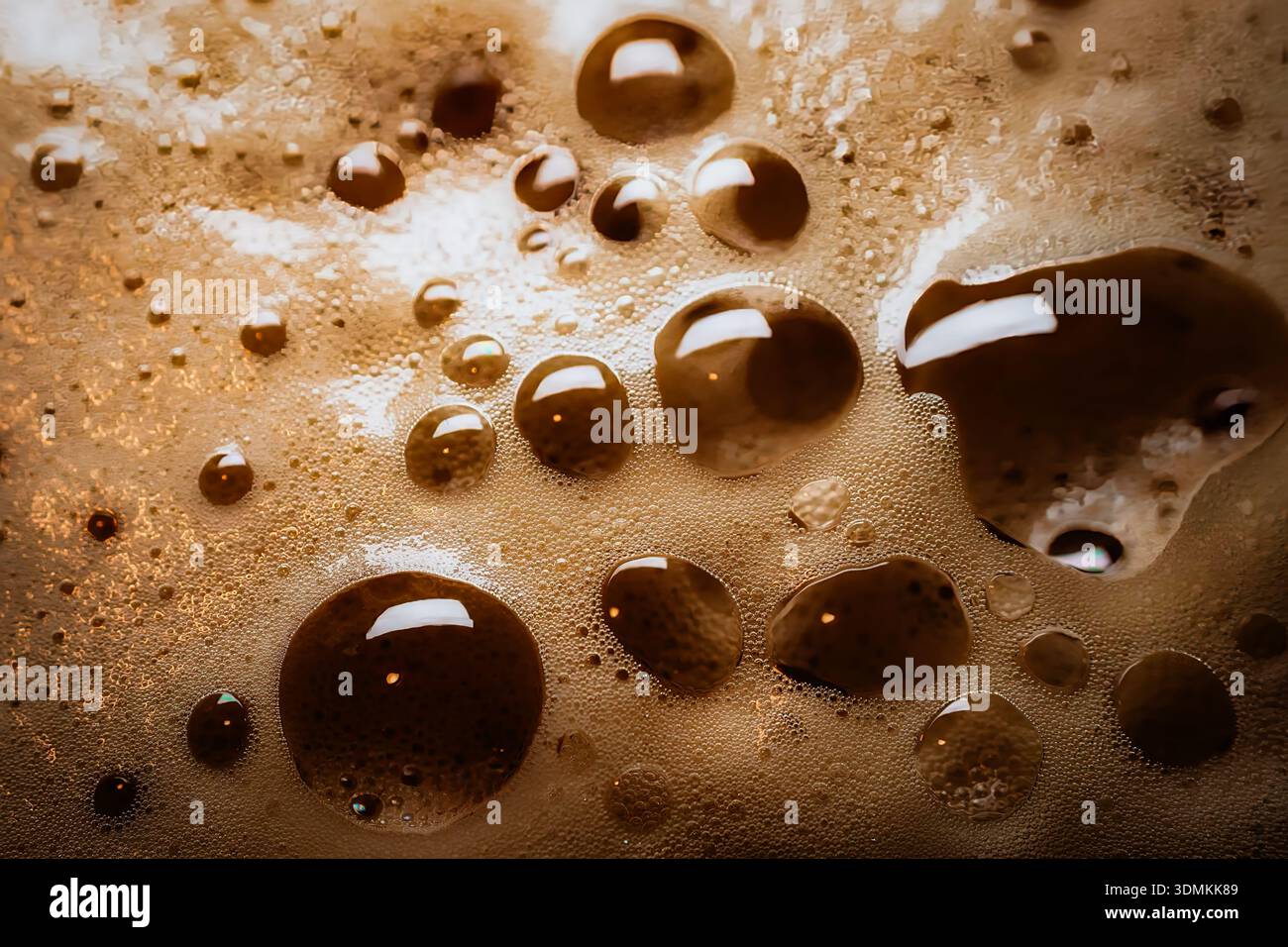 Gros plan de mousse pour boissons gazeuses avec bulles texturées montrant des tons bruns riches et des reflets brillants sous une lumière ambiante chaude. Banque D'Images