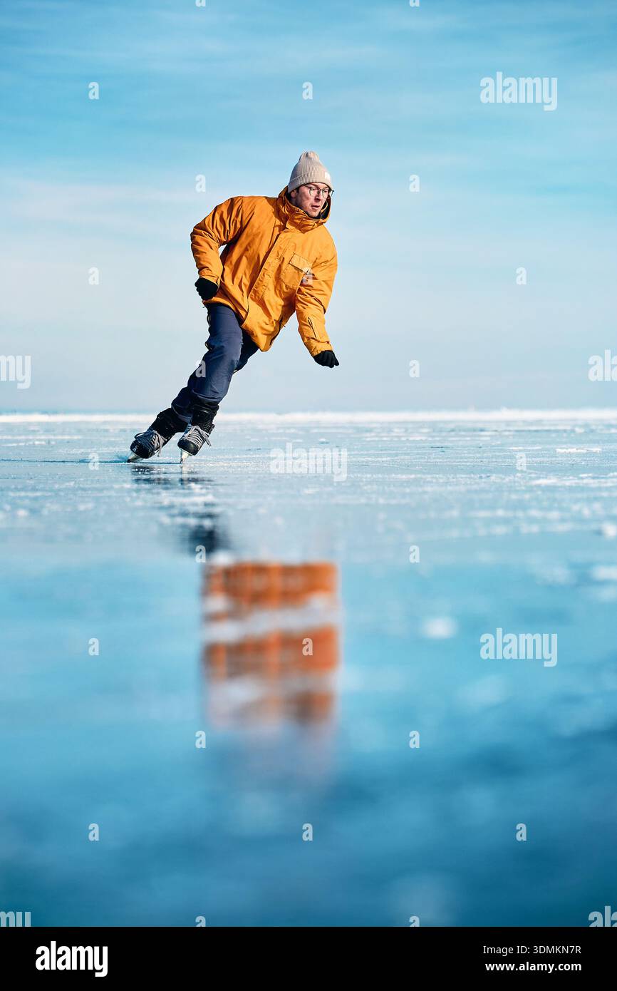 Homme athlète en veste jaune patin sur glace sur le lac gelé avec des fissures et de la neige à l'heure d'hiver Banque D'Images