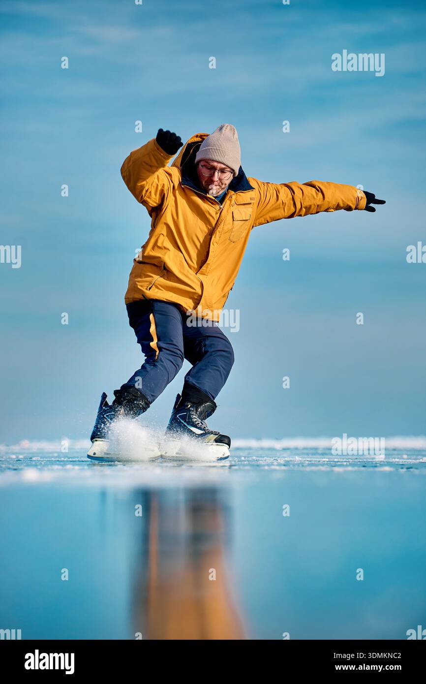 Homme athlète en veste jaune patin sur glace sur le lac gelé avec réflexion à l'heure d'hiver Banque D'Images