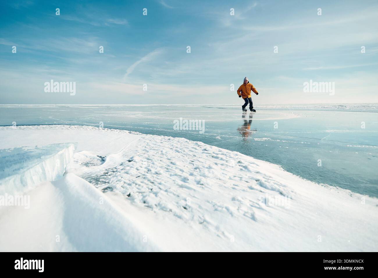 Homme athlète en veste jaune patin sur glace sur le lac gelé avec des fissures et de la neige à l'heure d'hiver Banque D'Images